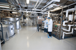 Two people in lab coats and hairnets inspect equipment in an industrial facility with metal tanks and an array of pipes.