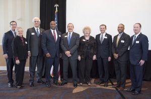 Nine people in formal attire stand together indoors, smiling. An American flag is visible in the background on the left side.