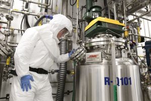 A person in protective gear examines industrial equipment in a complex facility filled with pipes and machinery.