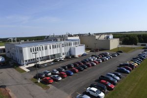 Modern industrial facility with multiple buildings, large parking lot full of cars, situated in a green rural landscape under a clear blue sky.