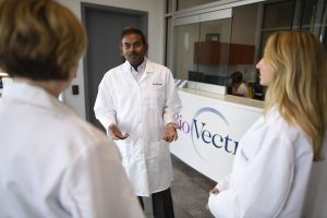Three people in lab coats converse near a reception area with the BioVectra logo visible in the background.