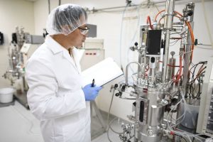 A person in lab coat and gloves examines equipment in a laboratory, holding a notebook. Complex machinery with tubes surrounds the workspace.
