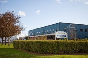 A blue industrial building with "BioVectra" signage, surrounded by green hedges and trees, under a clear sky. No people visible.