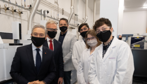 Five individuals wearing masks and lab coats pose in a laboratory with scientific equipment and industrial piping visible in the background.