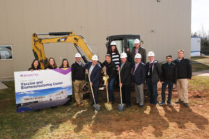 A group of eleven people in hard hats participate in a groundbreaking ceremony at a future vaccine and biomanufacturing center site.