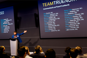 A person presents at a conference. A screen displays various Canadian companies' locations. Audience members watch attentively in a well-lit room.
