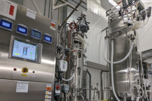 A spacious industrial lab with large stainless steel tanks and complex control panels featuring digital displays and connecting tubes.