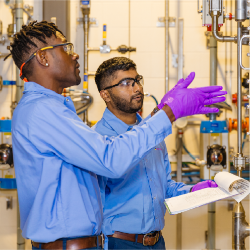 Two people wearing safety goggles and blue shirts discuss equipment in a laboratory setting. One person gestures while holding a booklet, both attentive.