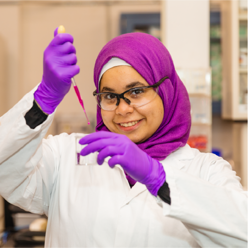 A person in a lab coat, purple gloves, and glasses uses a pipette in a laboratory setting, demonstrating scientific experimentation.