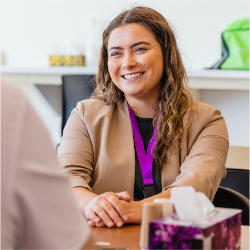 A person with a purple lanyard smiles at another person across a table with tissues and a brightly colored bag in the background.