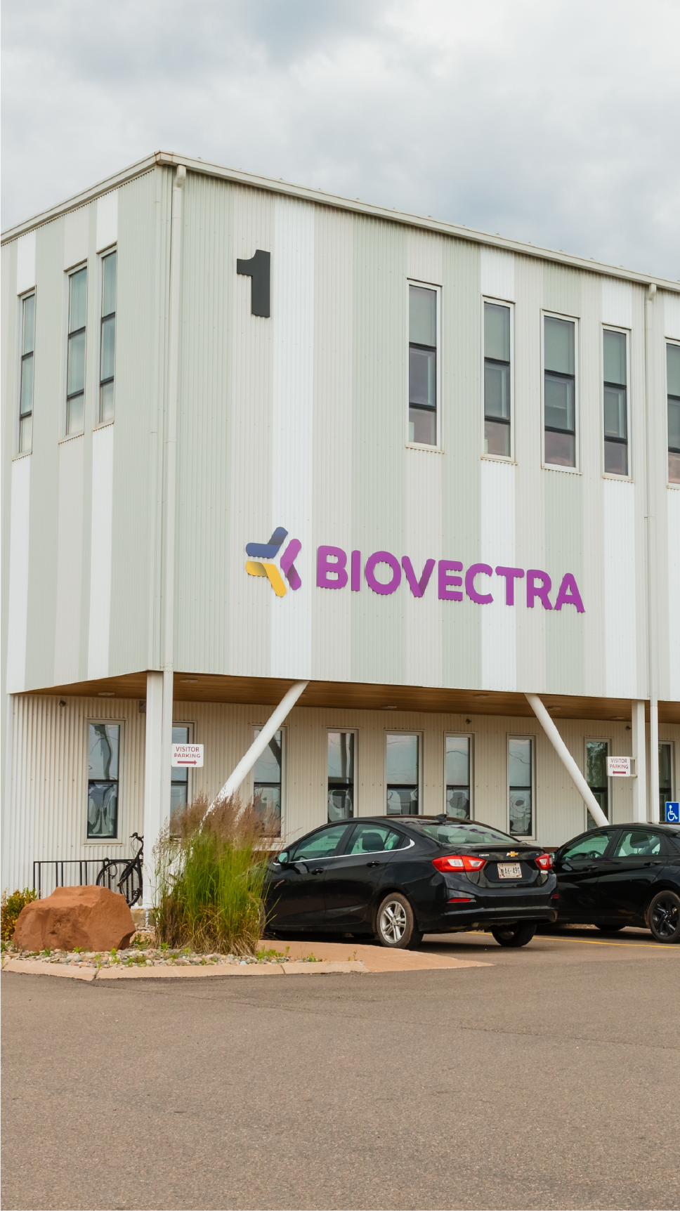 A modern Biovectra building with large windows and parked cars outside. Overcast sky above industrial-looking structure with signage and minimal landscaping.
