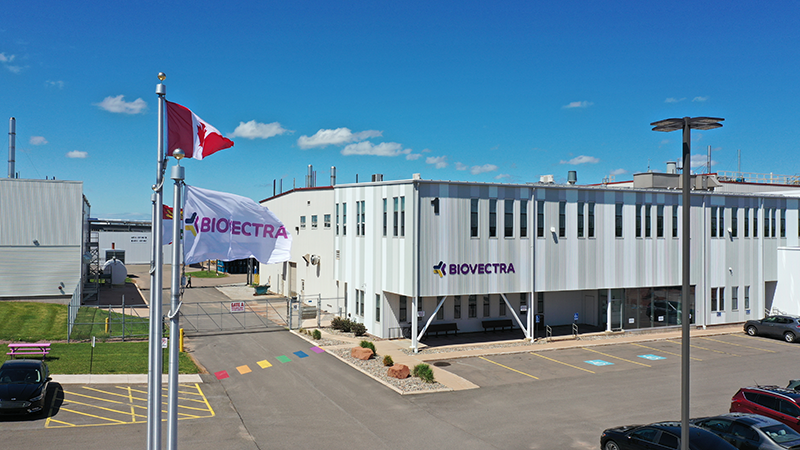 Modern industrial building with "BIOVECTRA" signage. Two flags, including Canadian, are visible. Parking area and clear blue sky in the background.