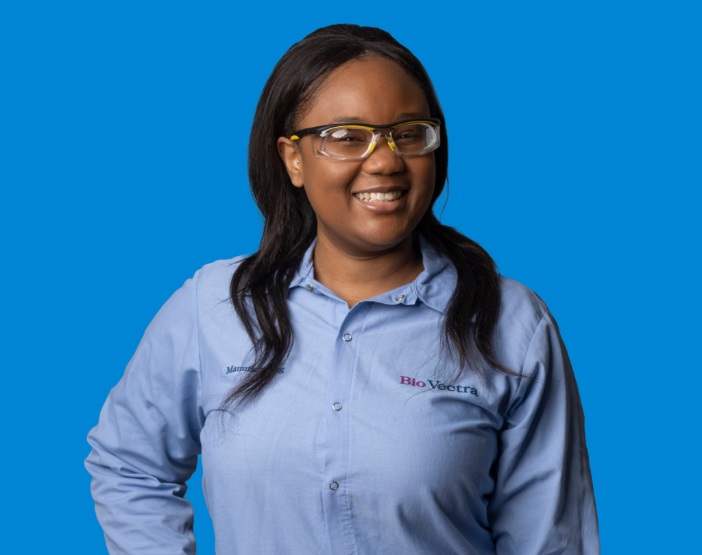 A smiling person in safety glasses and blue shirt against a purple background. No recognizable landmarks or historical buildings are present.