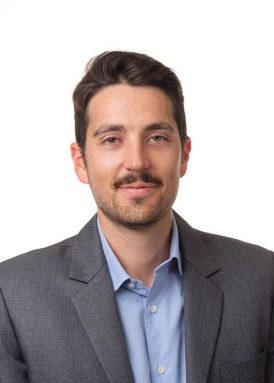 A person with short hair and a mustache wearing a gray suit and blue shirt, smiling against a plain white background.