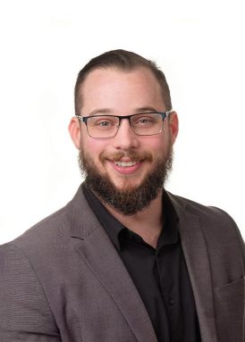 A person with glasses and a beard smiles, wearing a suit jacket over a black shirt, against a plain white background.