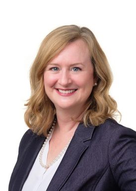 A person with shoulder-length blonde hair smiles, wearing a dark blazer, white blouse, and pearl necklace. Plain white background.