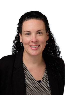 A person with curly hair is smiling, wearing a patterned shirt and dark blazer against a plain white background.