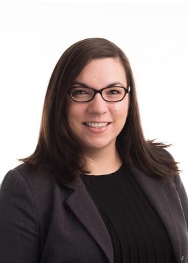 A person wearing glasses with dark hair smiles, dressed in a professional black top and blazer, on a plain white background.