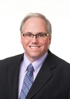 A person wearing glasses, a suit, and a patterned tie smiles at the camera against a plain white background.