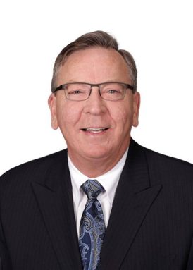 A person wearing glasses and a suit smiles against a plain white background. No recognizable landmarks or historical buildings are visible.