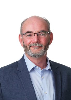 A person with glasses and a beard, wearing a suit and light blue shirt, smiles against a plain white background.