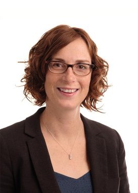 A person with short curly hair and glasses, wearing a black blazer and blue shirt, smiles against a plain white background.